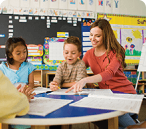 teacher sitting at desk with two students pointing at paper and
                        smiling